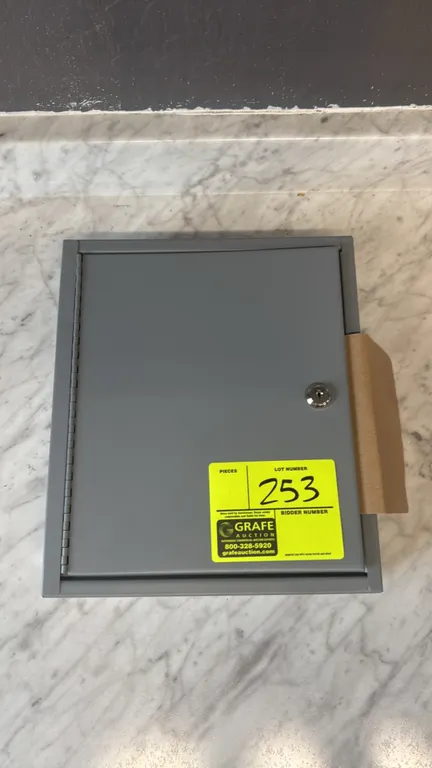 Metal key storage box. Features single door with cylinder lock, interior slotted key hook racks, and includes key tags. Gray metal construction. 10 1/