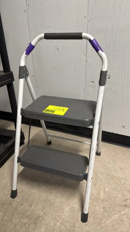 Two-step folding metal step stool. Features a white tubular metal frame, slip-resistant textured steps, top hand grip, and rubber feet.