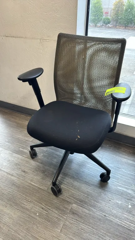Office chair. Features a mesh back, black upholstered fabric cushioned seat, and armrests. Mounted on a 5-star base with dual-wheel casters.