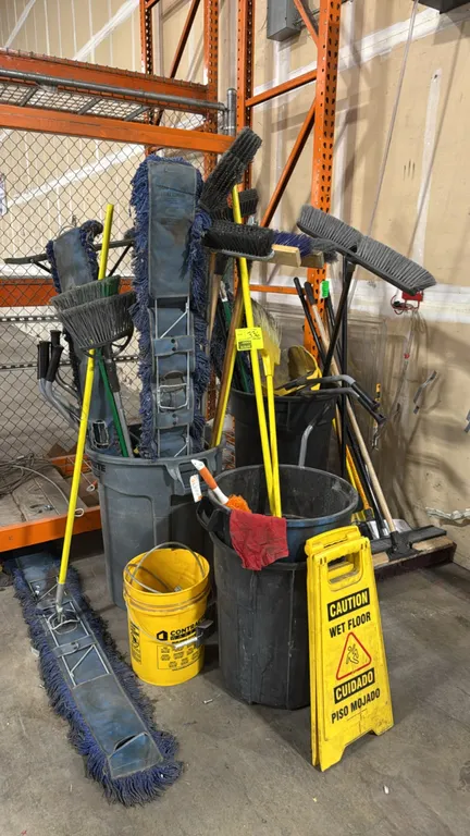 Janitorial and maintenance supply lot. Includes commercial trash cans, push brooms, angle brooms, wide dust mops, upright dustpans, folding 'Caution W