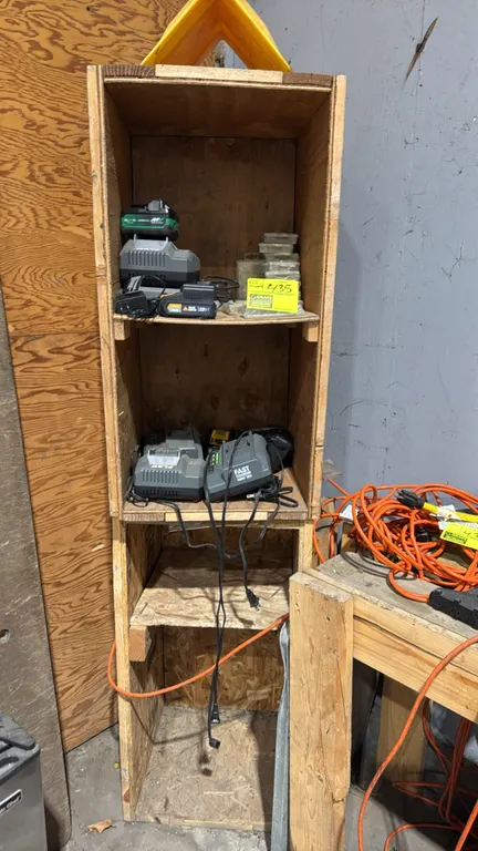 Plywood storage shelving unit with assorted power tool battery chargers and hardware. Includes Flex, DeWalt, and Blue Ridge chargers, battery packs, a