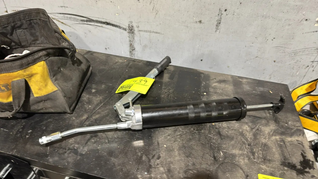 Lever-action metal grease gun. Features a lever handle with textured grip, rigid extension tube, coupler nozzle, and rear T-handle plunger. Tool bag a