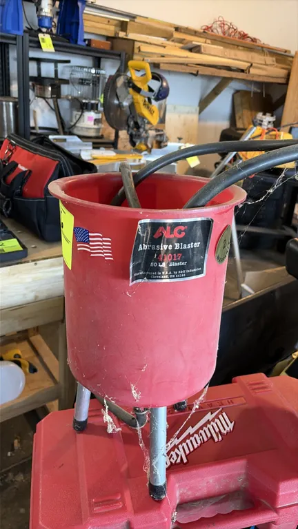 ALC 41017 abrasive blaster. Features 50 lb capacity hopper, blast hose, and blast gun. Mounted on tripod legs.
