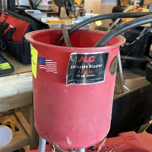ALC 41017 abrasive blaster. Features 50 lb capacity hopper, blast hose, and blast gun. Mounted on tripod legs.