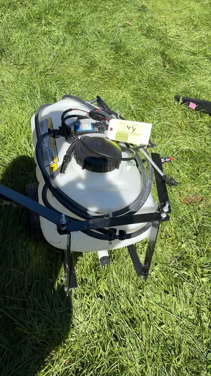 Brinly 12-gallon pull-behind boom sprayer. Features a translucent polyethylene tank, rear spray boom, hand spray wand with hose, and an Everflo 12V el