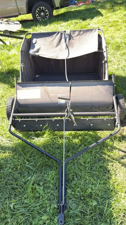 Brinly tow-behind lawn sweeper. Features rotary sweep brushes, de-thatcher tines, height adjustment lever, and collection hopper with dump rope. Equip