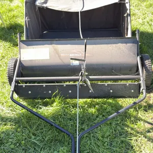 Brinly tow-behind lawn sweeper. Features rotary sweep brushes, de-thatcher tines, height adjustment lever, and collection hopper with dump rope. Equip