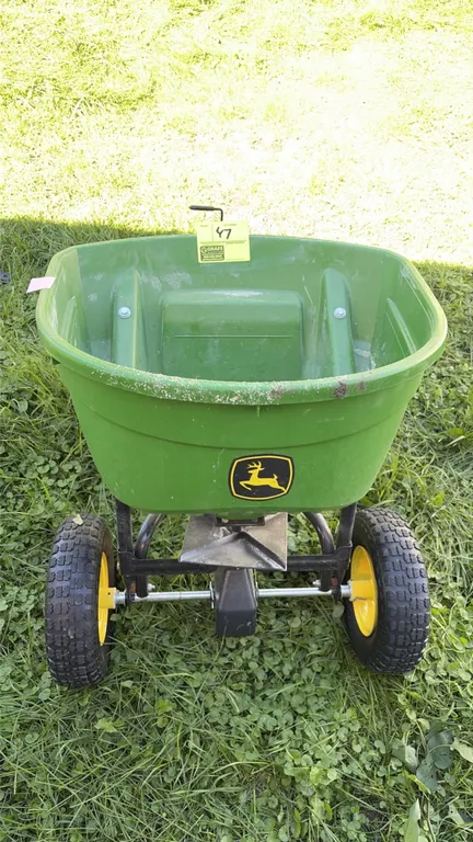 John Deere tow-behind lawn seeder and broadcast spreader. Features green poly hopper, flow control lever, broadcast spreader plate, and pneumatic tire