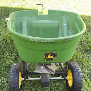 John Deere tow-behind lawn seeder and broadcast spreader. Features green poly hopper, flow control lever, broadcast spreader plate, and pneumatic tire