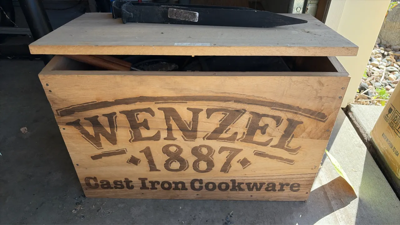 Wenzel 1887 cast iron cookware set with wooden storage chest. Includes a Dutch oven with lid, nested skillets, camp pie iron, metal stakes, and zipper