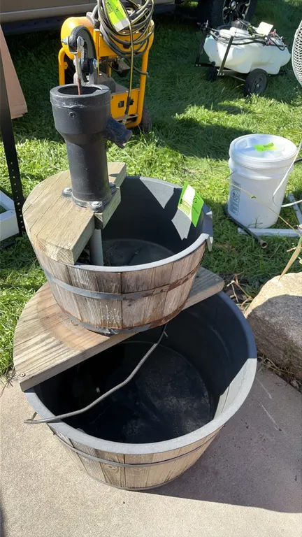 Rustic two-tier barrel water fountain lawn decor. Features a decorative cast metal hand well pump, tiered faux-wood barrels with plastic interior line