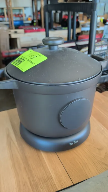 Our Place electric rice cooker. Features a matte charcoal exterior, matching lid with top knob, integrated side handles, and circular front display. M