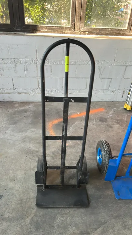 Metal 2-wheel hand truck. Features a tubular metal frame, loop handle, integrated wheel guards, and (2) solid rubber tires. Additional hand trucks and