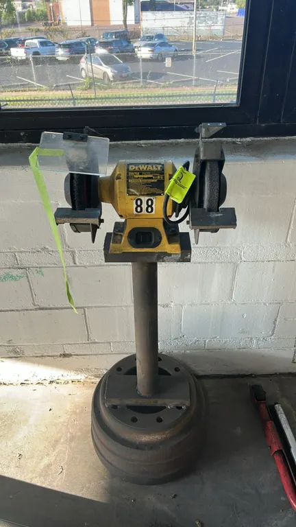 DeWalt DW758 8" bench grinder. Features adjustable tool rests, left clear eye shield, grinding wheel, and wire wheel. Mounted on a steel pedestal stan