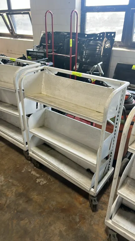 Metal portable multi-tier shelving cart. Features 3 sloped metal shelves with integrated backstops and white finish. Dimensions: 32" x 16". Mounted on