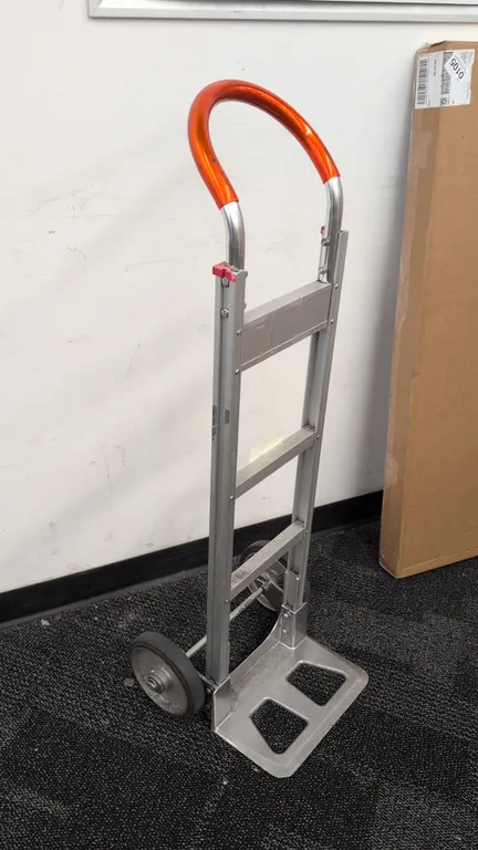 Win-Holt aluminum hand truck. Features a curved loop handle with orange grip, aluminum frame with three cross braces, and open die-cast nose plate. Mo