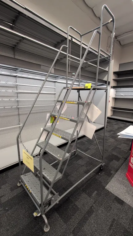 Tri-Arc mobile metal stock ladder. Features 7 steps with perforated slip-resistant treads, top platform, tubular handrails, and upper guardrails. Dime