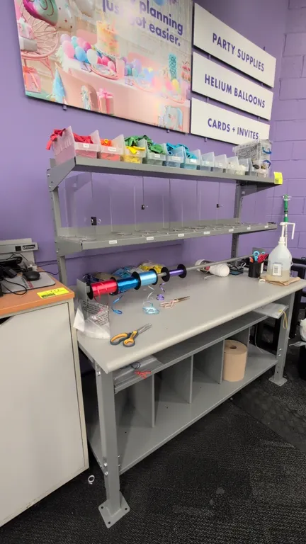 Laminate adjustable workstation table. Features gray metal frame, two-tier upper shelf with clear dividers, roll dispenser rod, and lower divided stor