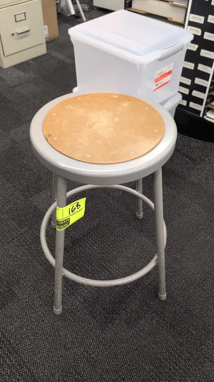 Metal shop stool. Features a round hardboard seat insert, gray painted steel frame, and circular footrest ring. 24"H. Mounted on four legs with protec