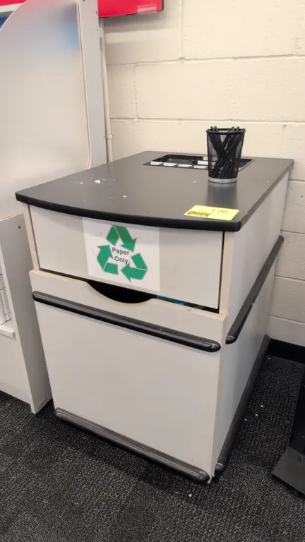 Laminate waste bin holder cabinet. Features a black laminate top with recessed supply tray, front cutout opening, single hinged lower door, and black