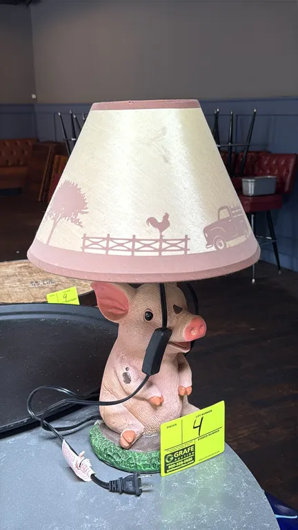 Tabletop pig-themed accent lamp. Features a cast resin seated pig figurine base and a tapered shade with a printed country farm motif depicting a pick