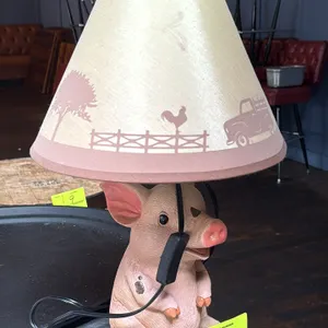 Tabletop pig-themed accent lamp. Features a cast resin seated pig figurine base and a tapered shade with a printed country farm motif depicting a pick