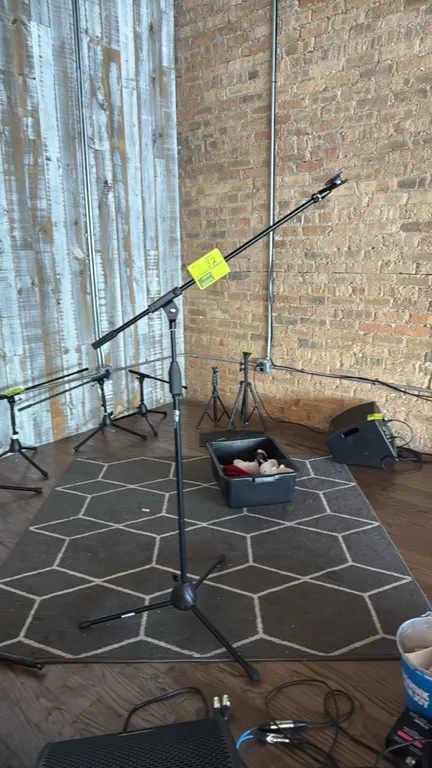 Boom microphone stand. Features an adjustable boom arm, height adjustment clutch, and attached microphone clip. Mounted on a folding tripod base.
