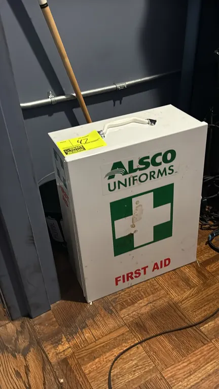 Alsco Uniforms metal first aid kit cabinet. Features a hinged door, top carrying handle, and interior shelving. Includes assorted first aid supplies.