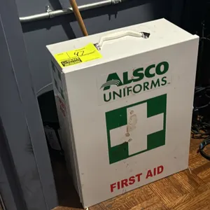 Alsco Uniforms metal first aid kit cabinet. Features a hinged door, top carrying handle, and interior shelving. Includes assorted first aid supplies. 
