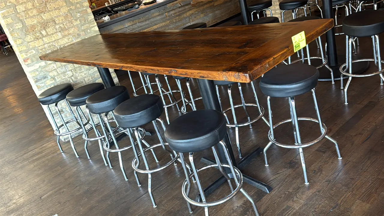 Wood bar height table with (10) black padded swivel stools. Table features a stained wood top mounted on dual black metal pedestal cross bases. Stools