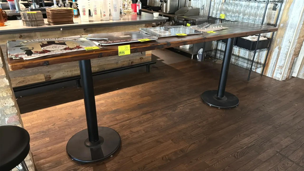 Wood bar-height table. Features a plank-style wood tabletop. Dimensions: 120"L x 24"W x 42.5"H. Mounted on dual black metal pedestal column bases with