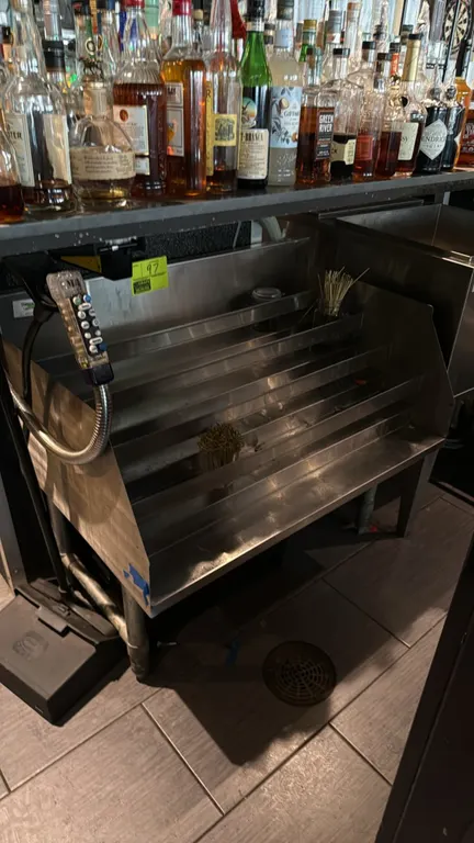 Stainless steel underbar liquor bottle display cabinet. Features a 5-tier stepped bottle rack design with bottle guardrails and an integrated backspla