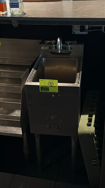 Stainless steel underbar hand sink. Features single sink compartment, integrated backsplash, splash-mount faucet with dual lever handles, and enclosed