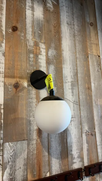 Round globe wall sconce light. Features a matte black curved arm, circular wall mounting plate, and white frosted globe shade. Hardwired wall mount. N