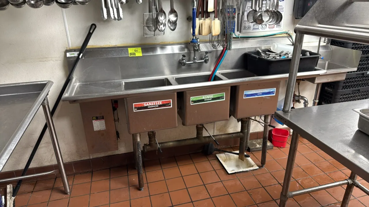 Stainless steel 3-compartment sink with left and right drainboards. Features splash-mounted pre-rinse faucet with flexible spray hose, lever drain val