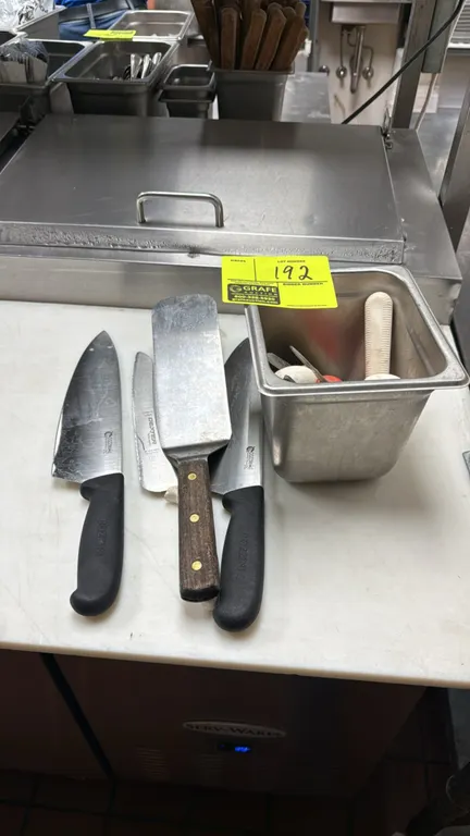 Assorted commercial kitchen cutlery and utensils. Includes Cozzini chef knives, Dexter serrated knife, wood-handled grill turner, and assorted oyster