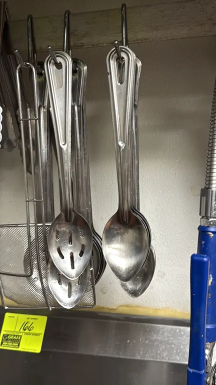 Assorted stainless steel serving spoons. Includes solid and slotted basting and serving spoons with grooved handles and hanging holes. Racks, hooks, a