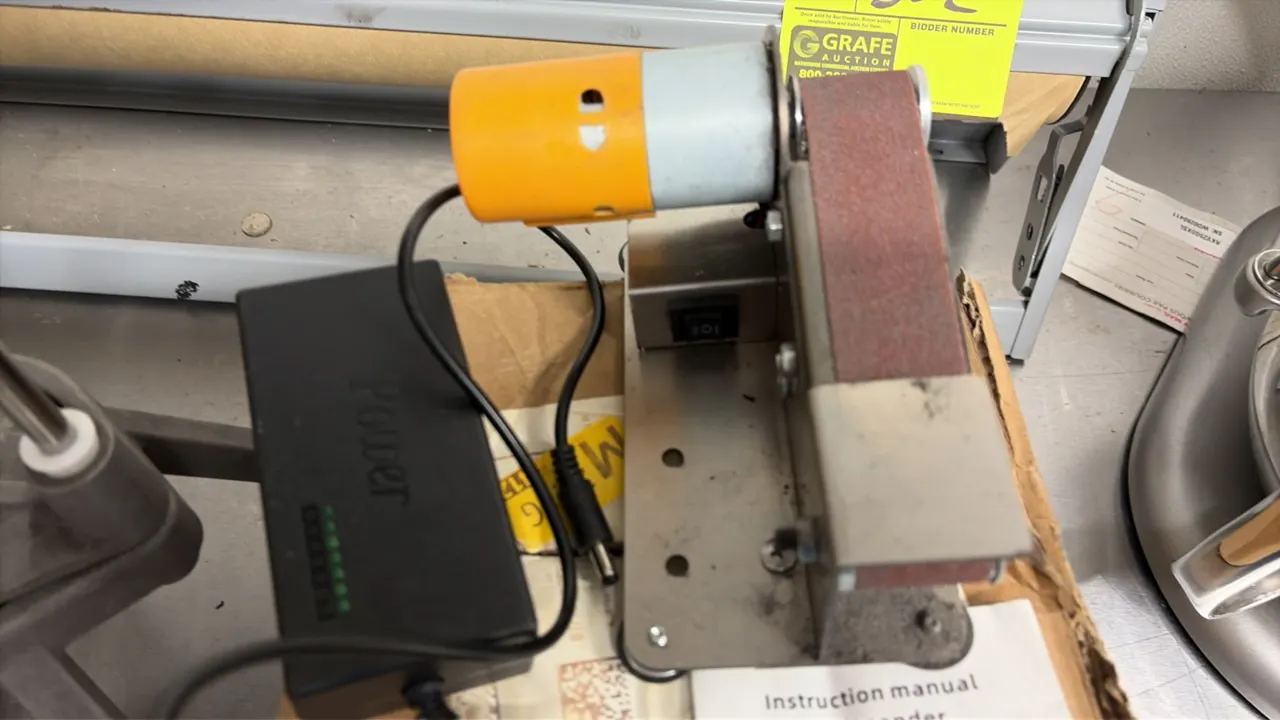 Mini electric benchtop belt sander. Features a metal frame, rubber foot pads, integrated rocker switch, and installed sanding belt. Includes external