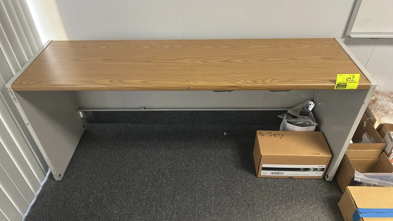 Office desk. Features a woodgrain laminate top and modesty panel. Mounted on a gray metal base. Dimensions: 70" x 20". Contents not included.