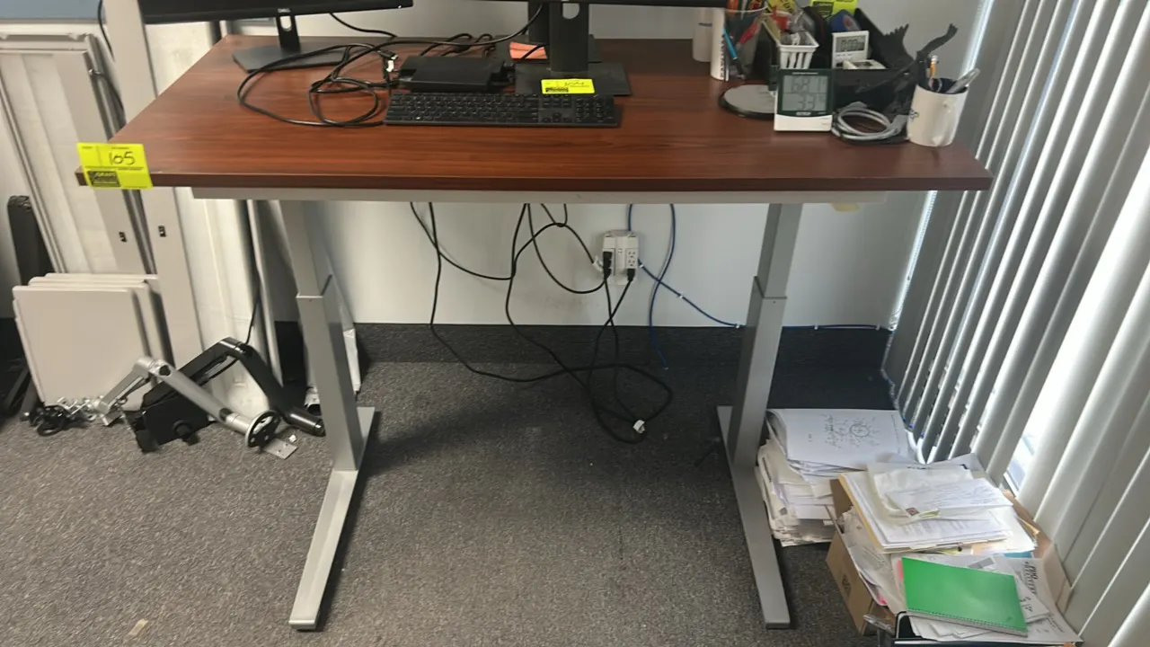 Adjustable height desk. Features a wood-tone laminate top and silver-tone metal base with T-legs. 60"W x 30"D. Monitors, electronics, and desktop cont