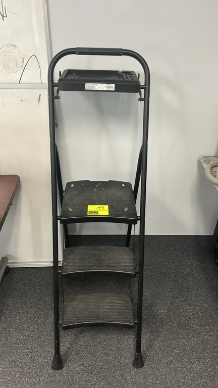 Folding 3-step ladder. Features black tubular metal frame, textured poly steps with large top platform, integrated top utility tray, and hand grip. Ru