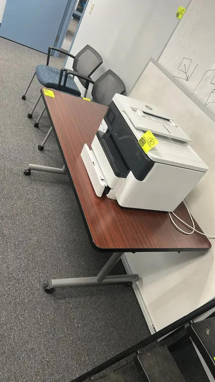 Portable laminate table. Features a woodgrain top with black edge banding. 60" x 30". Mounted on a silver-tone metal base with casters. Printer not in