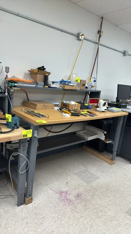 Adjustable-height laboratory work table. Features a composite wood top, upper metal riser shelf with integrated power strip, and lower metal utility s