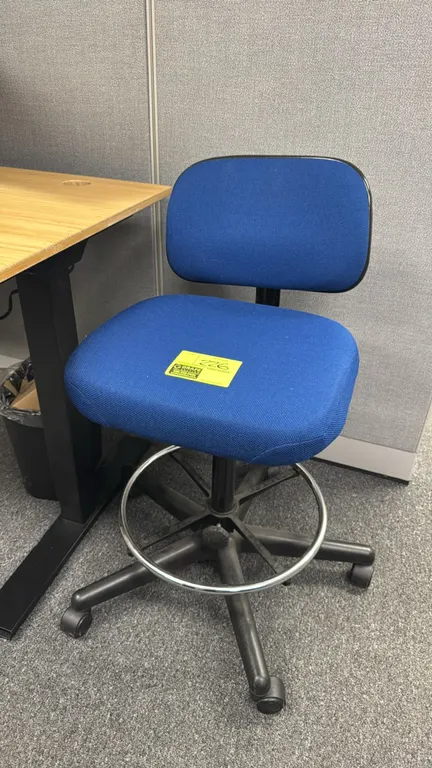 Upholstered laboratory drafting stool. Features blue fabric padded seat and back, height-adjustable backrest, and a chrome footrest ring. Mounted on a