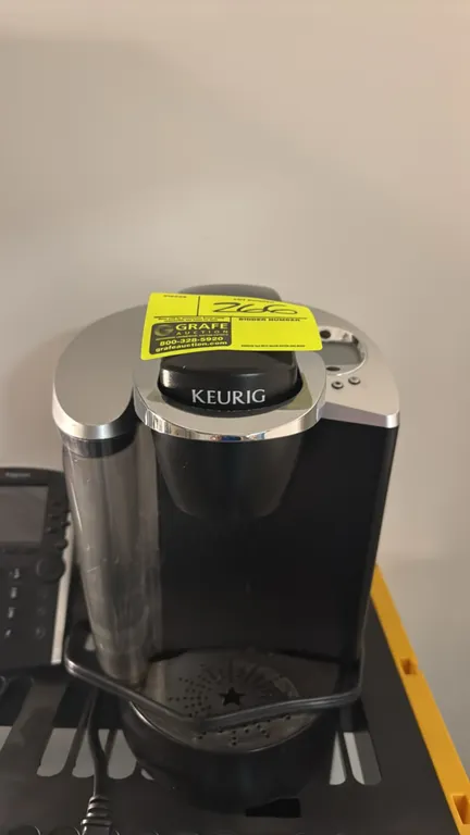Keurig single-serve coffee maker. Features side water reservoir, digital LCD display with push-button controls, chrome-accented brew handle, and remov