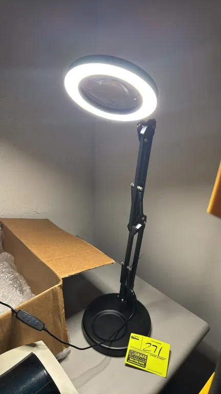 Adjustable LED magnifying task lamp. Features an integrated circular LED ring light with center magnifying glass, spring-assisted articulating arm, an