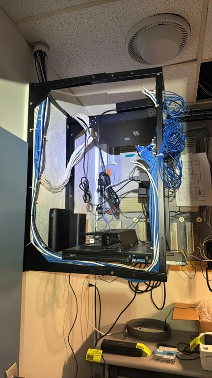 Wall-mounted IT server rack with contents. Includes APC battery backup UPS, SonicWall TZ600 firewall appliance, and Dell E05W network switch. Features