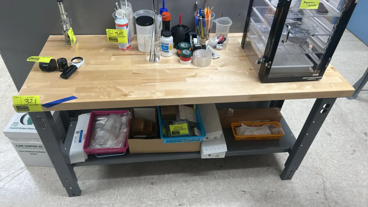 Two-tier workbench table. Features a wood butcher block top and metal lower shelf. Mounted on a painted steel base. Dimensions: 5' x 2'. Contents not