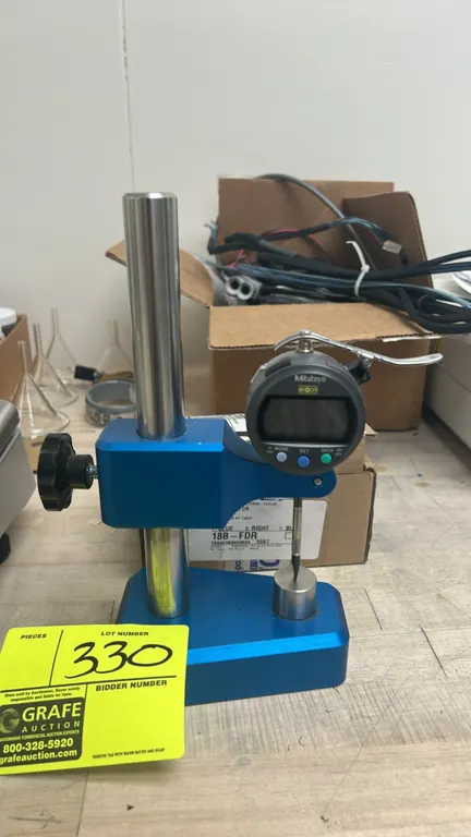 Mitutoyo digital thickness gauge. Features a digital indicator with lifting lever and a flat anvil. Mounted on an adjustable blue comparator stand.