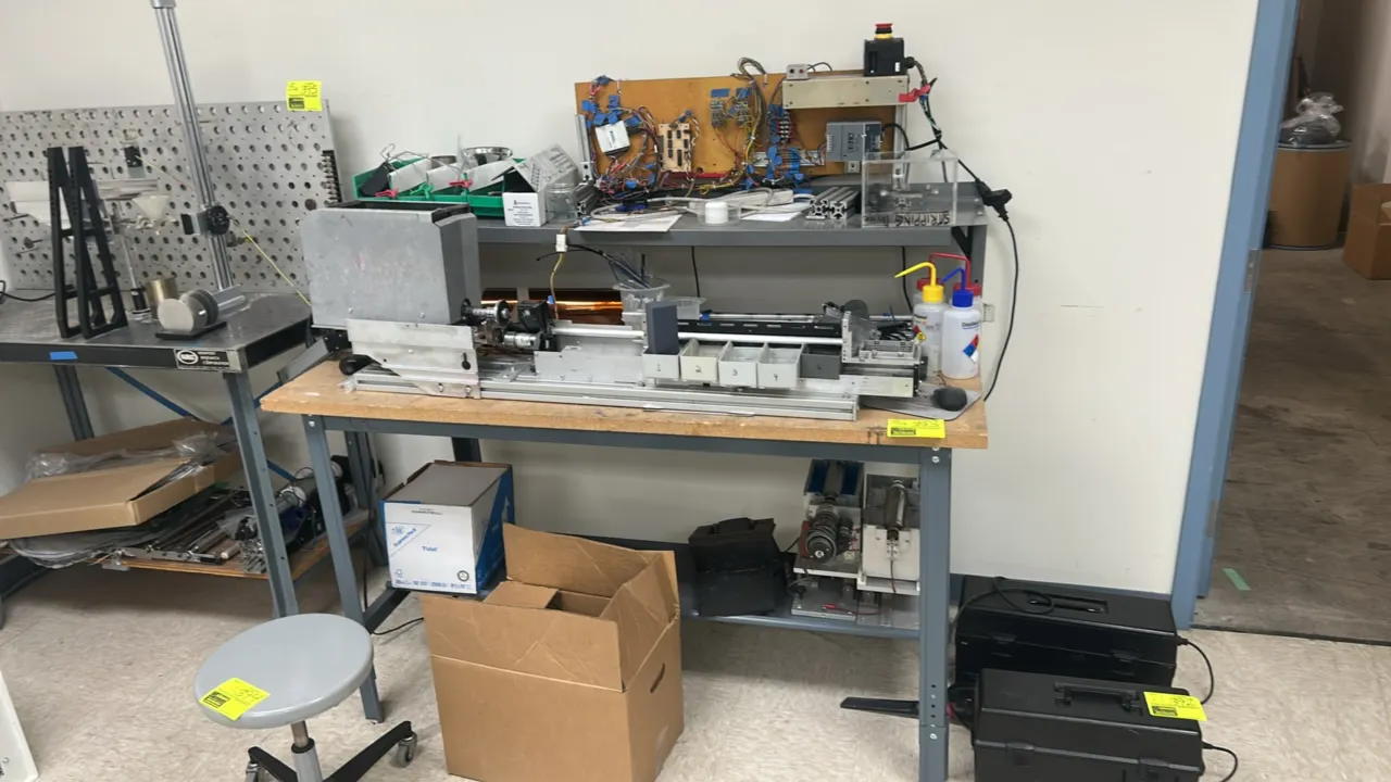 Metal workbench with custom industrial machinery. Features corkboard top, upper riser shelf, and lower shelf. Includes custom linear slide assembly, e